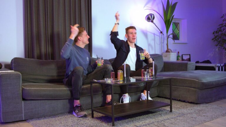 Twee vrienden op de bank vieren feest met kleurrijke cocktails op een glazen salontafel.