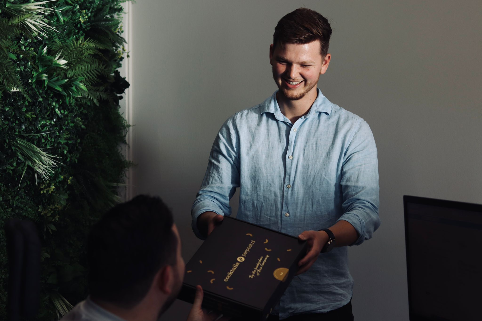 Man overhandigt een zwarte cocktailbox van Bar Company aan een gast met groene plantenwand op de achtergrond.