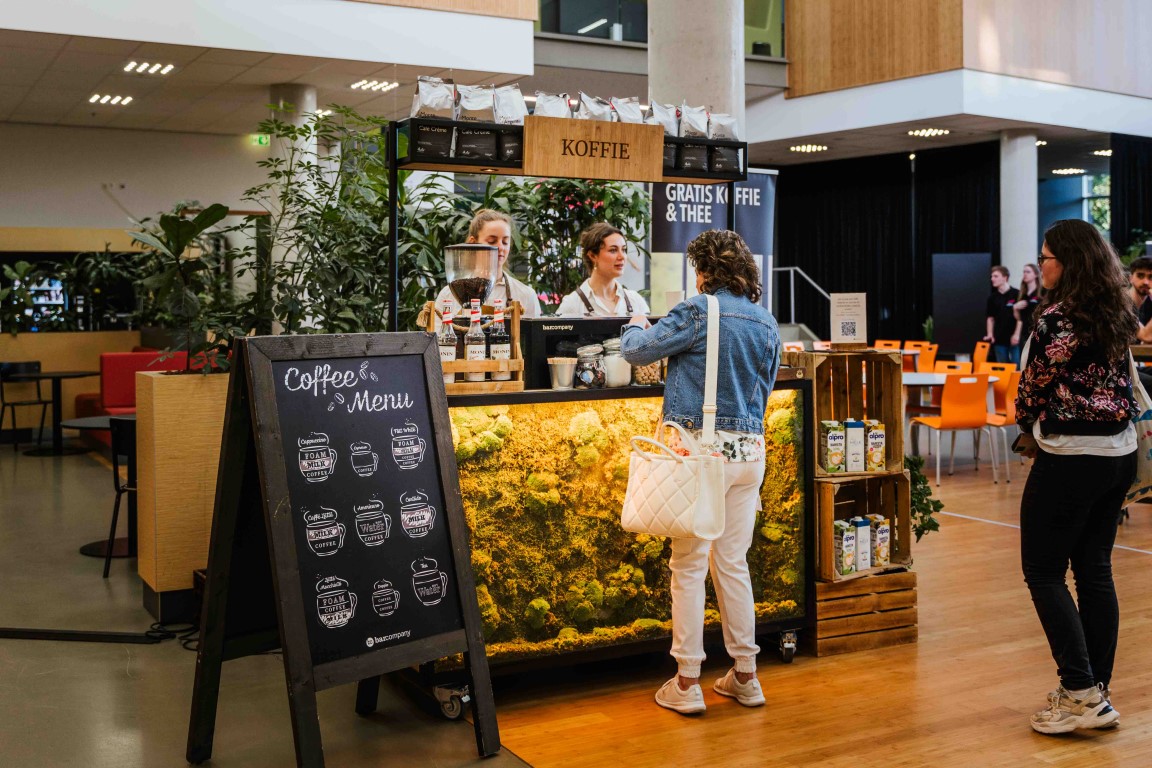 Twee barista's achter de groene mosbar met krijtbordmenu terwijl een gast bestelt.