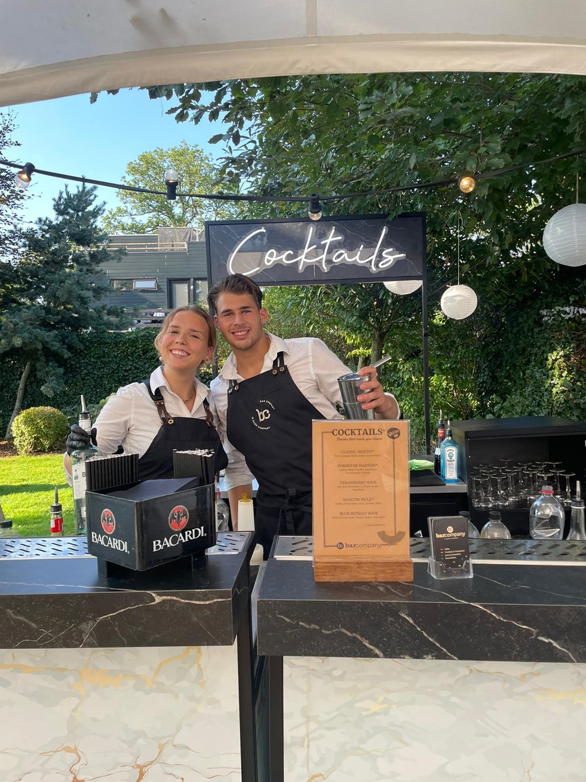 Twee vrolijke Bar Company bartenders achter een cocktailbar in de buitenlucht met een Cocktails-neonbord.