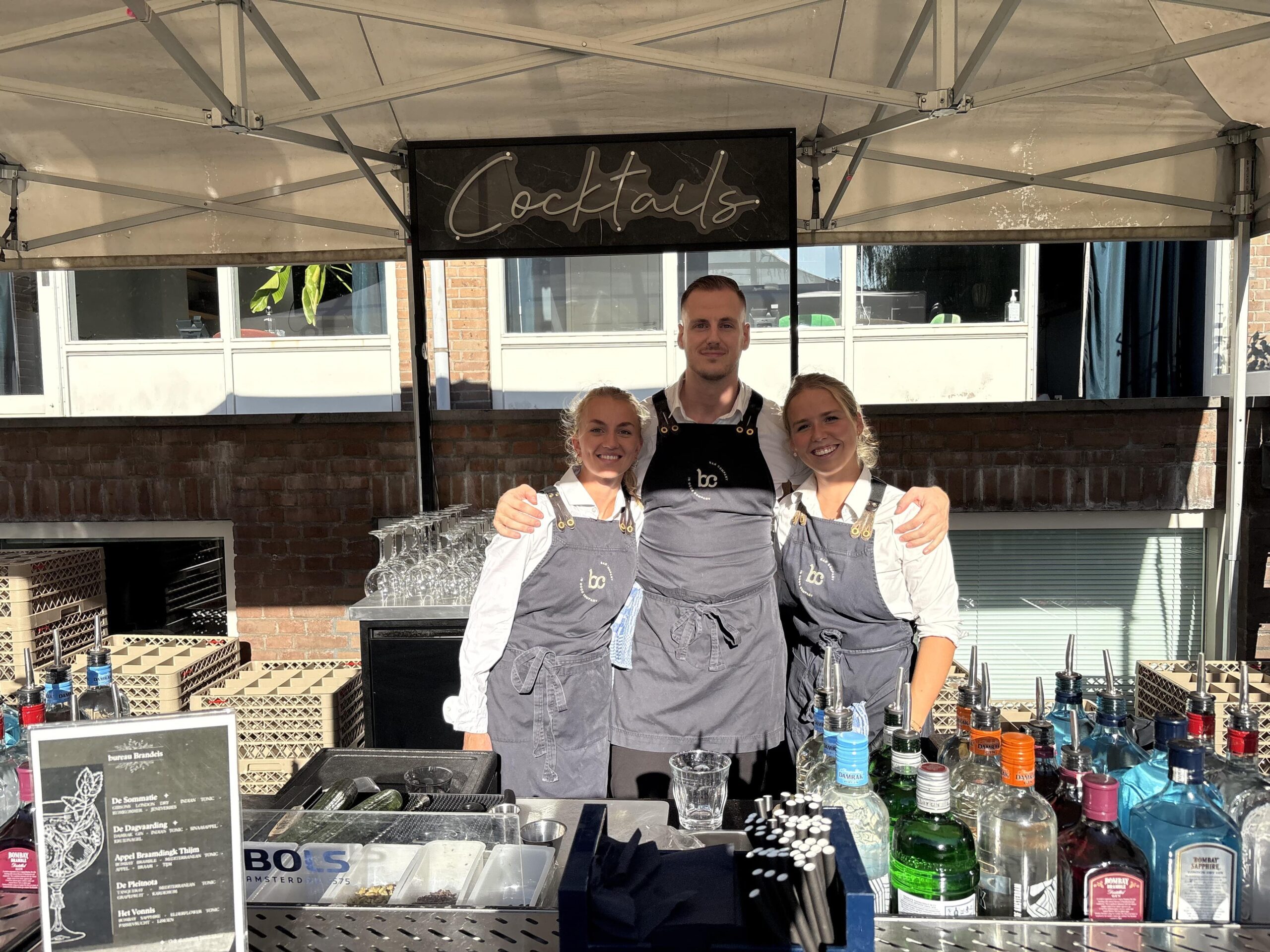 Drie Bar Company bartenders achter een cocktailbar op een zonnig buitenevenement met Bols en Bombay Sapphire