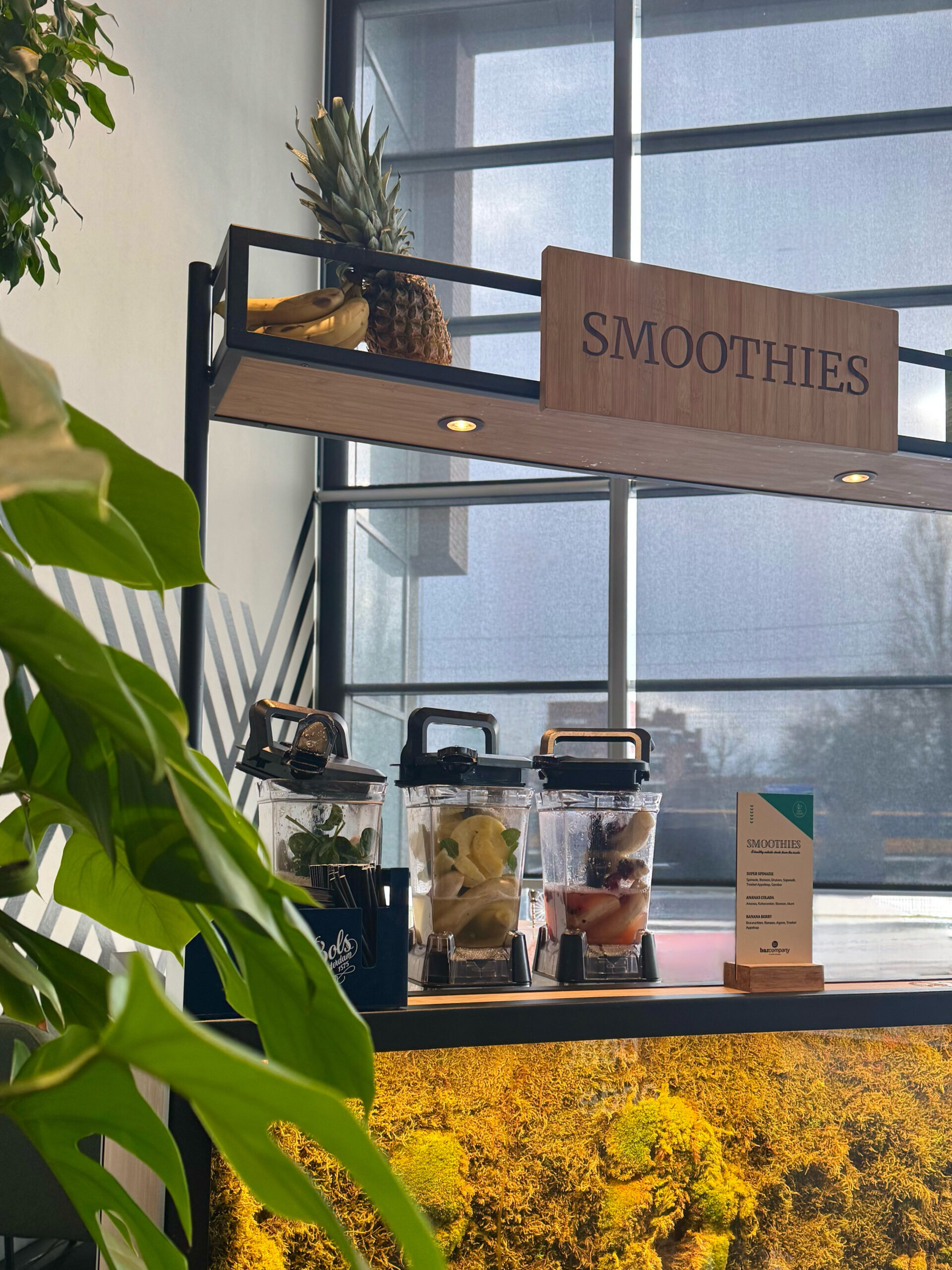 Bar met houten bord, verse ananas en banaan op de bovenplank en drie blenders klaargezet met fruit.