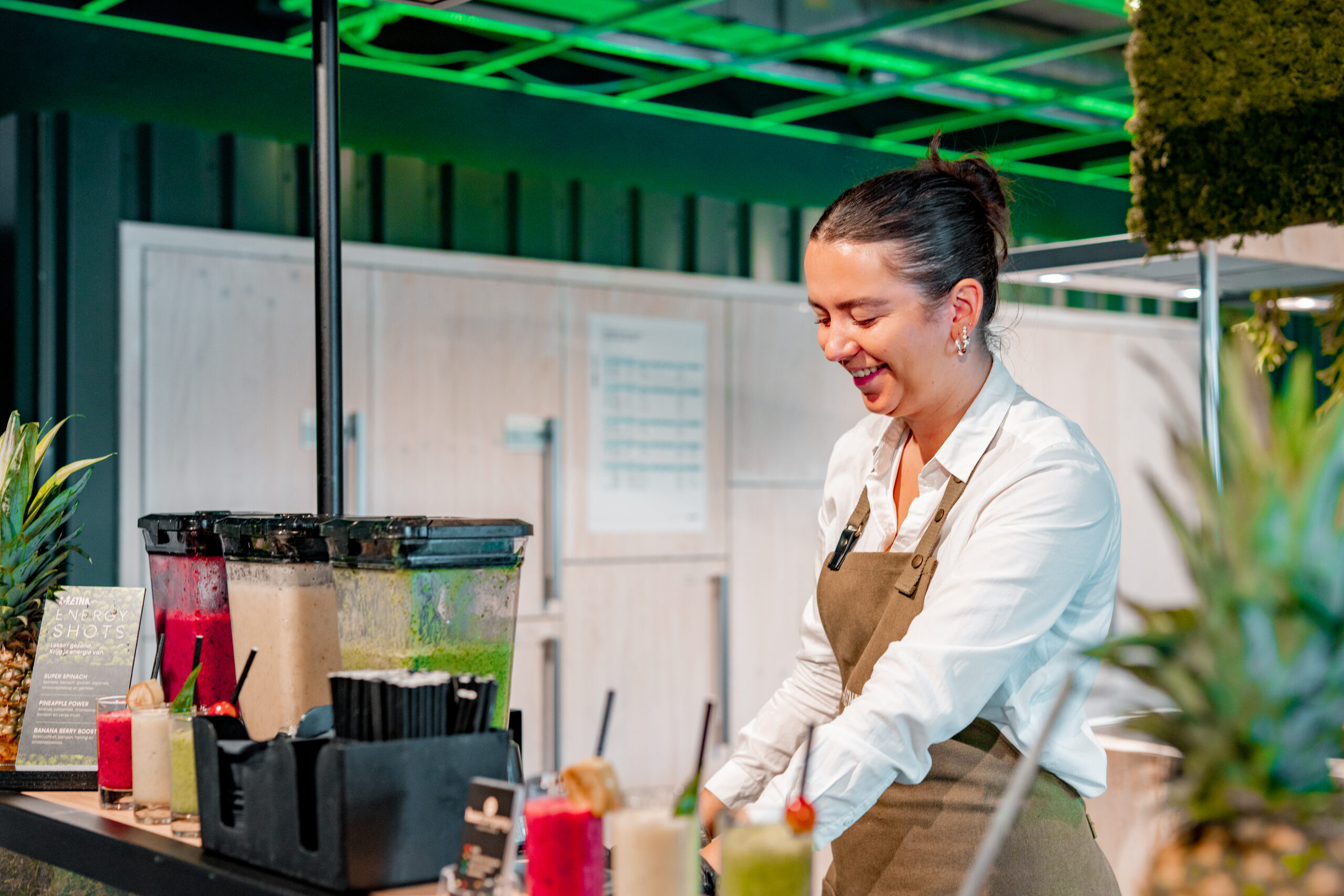 Bar Company medewerkster lacht achter een bar met drie blenders vol roze, beige en groene shakes.