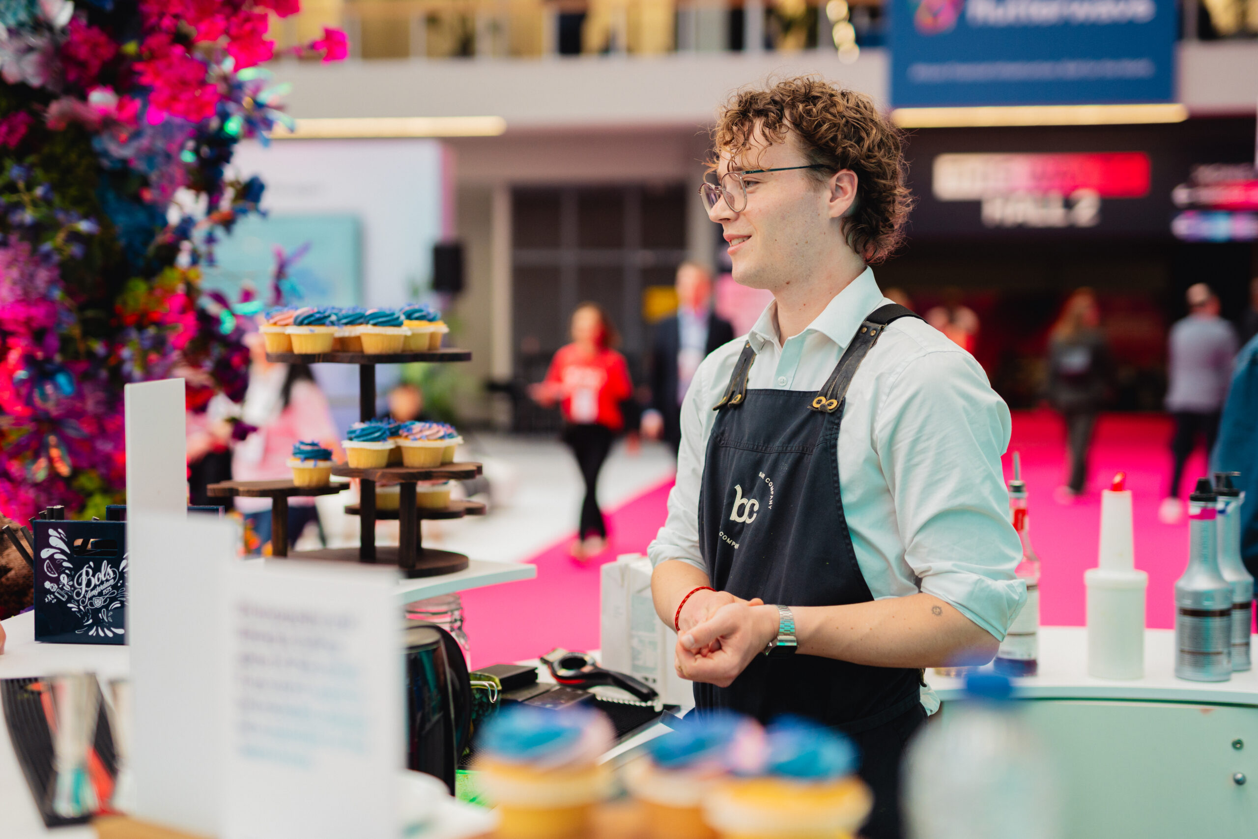 Barista van Bar Company achter de bar op een beursvloer, met cupcakes op houten etagères naast zich
