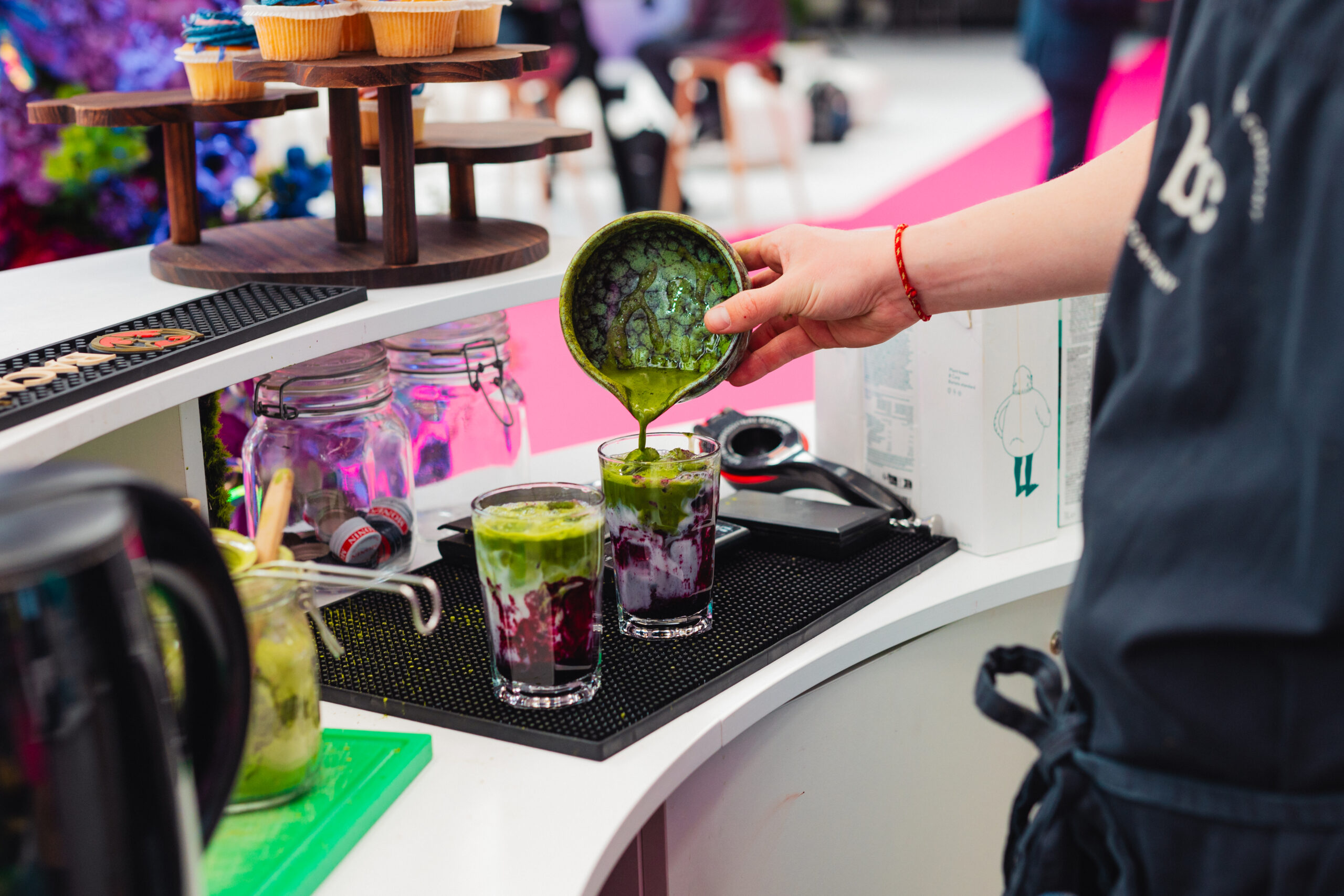 Barista schenkt opgeklopte matcha uit een keramische kom over de paarse ube-laag in twee glazen op de bar