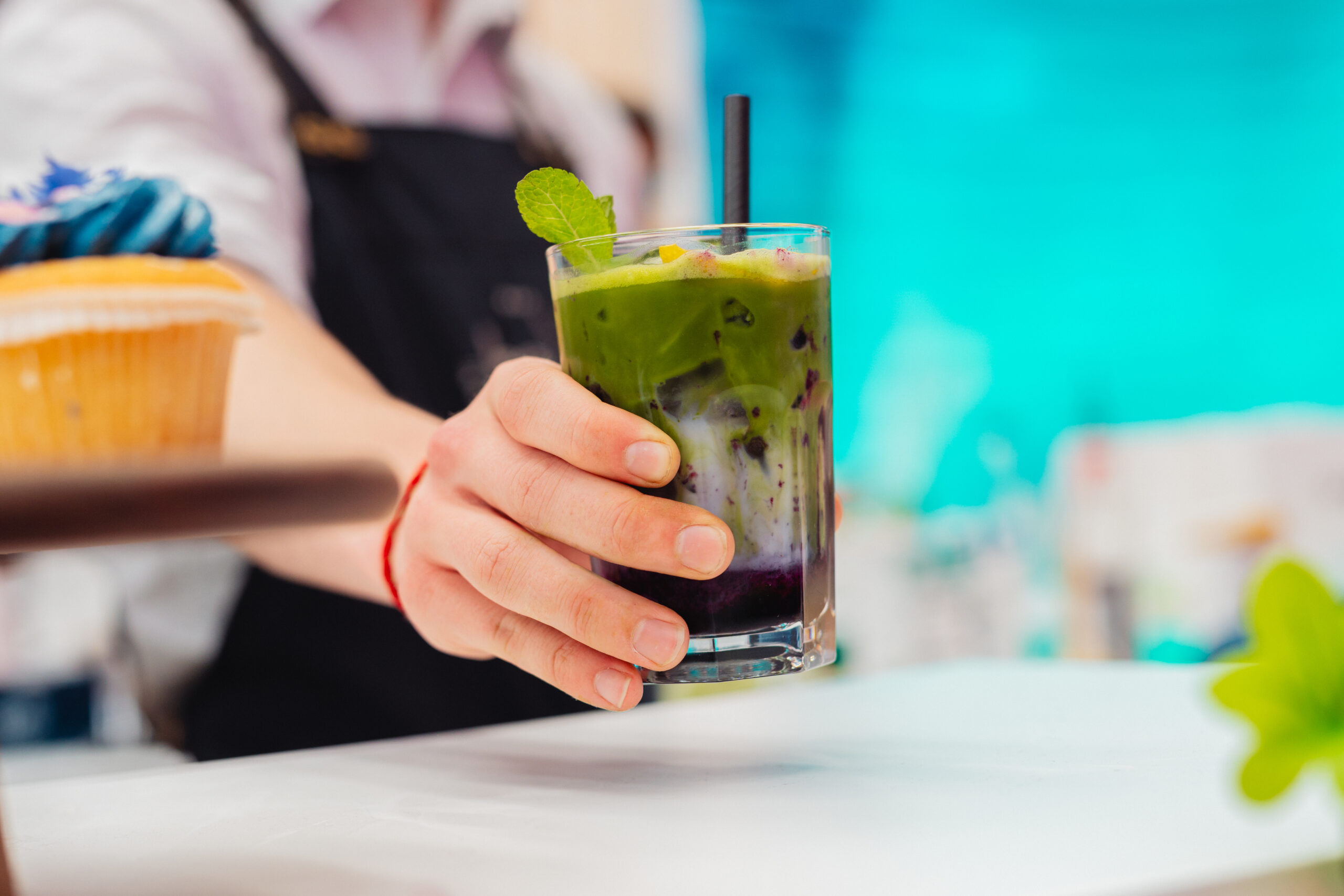 Bartender reikt een gelaagde groene shake met muntblaadje en rietje aan over de bar.