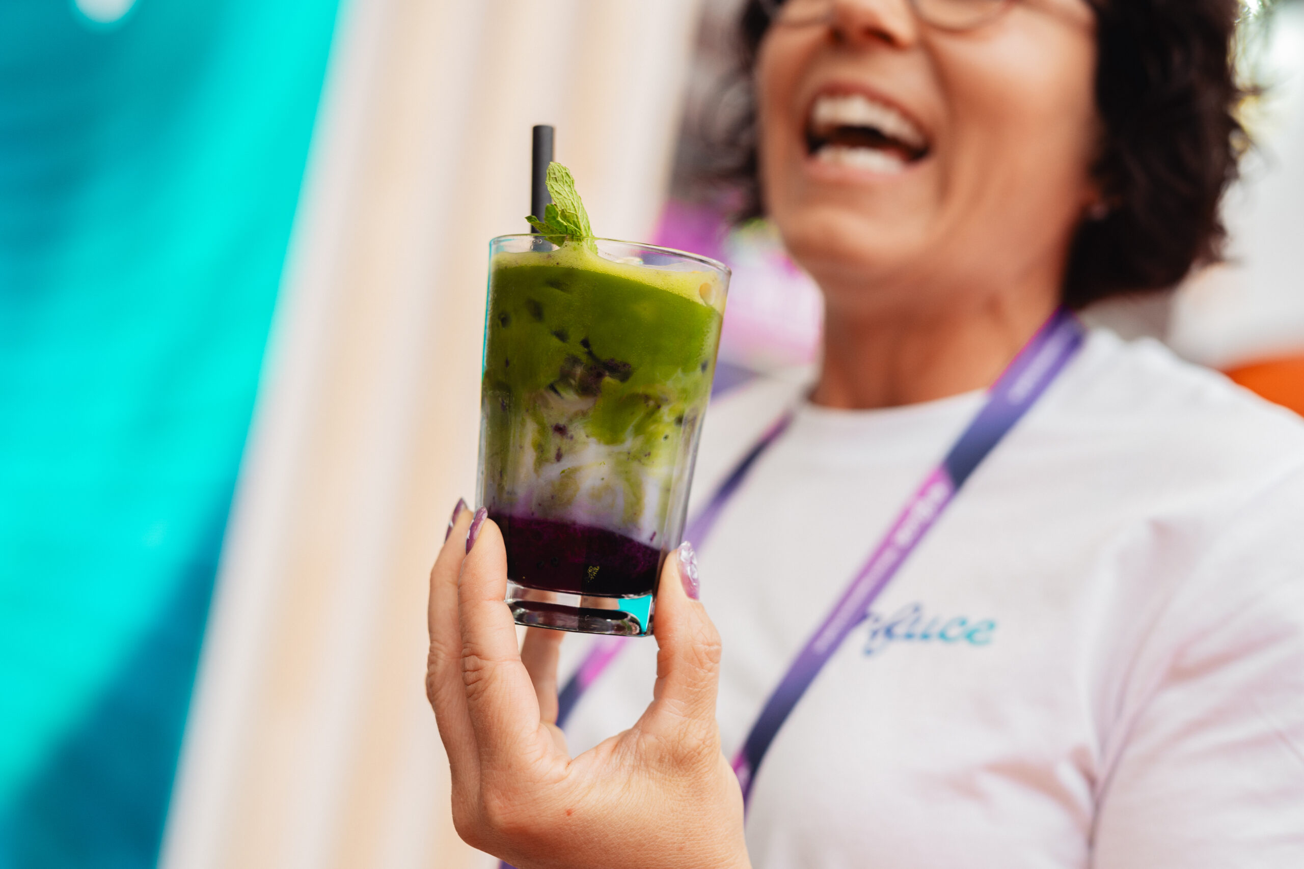 Lachende bezoekster houdt een glas ube matcha vast waarin de paarse, romige en groene laag onder elkaar zichtbaar zijn