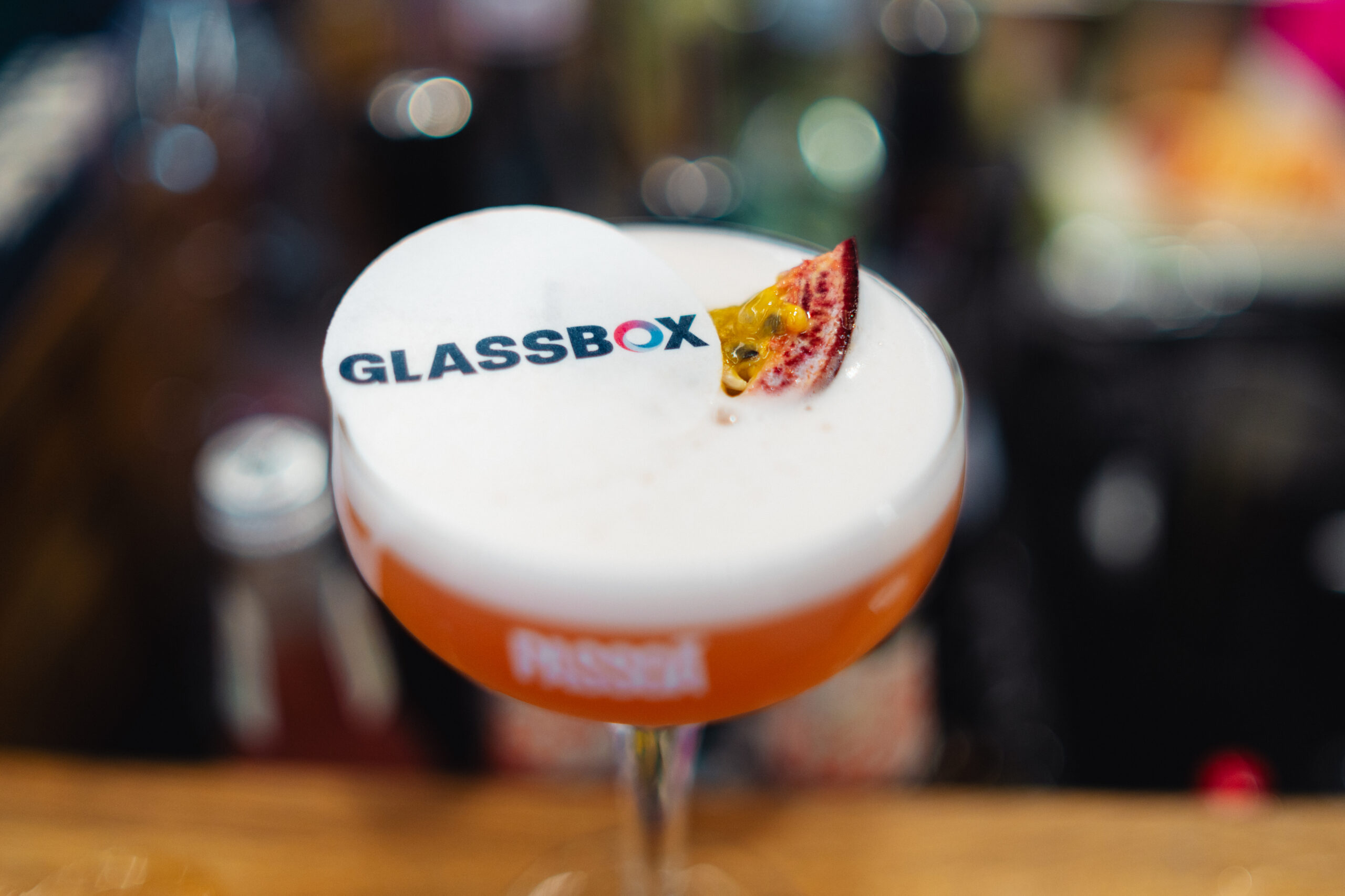 Oranje cocktail in een coupe met het logo van de klant in het schuim en een halve passievrucht als garnering