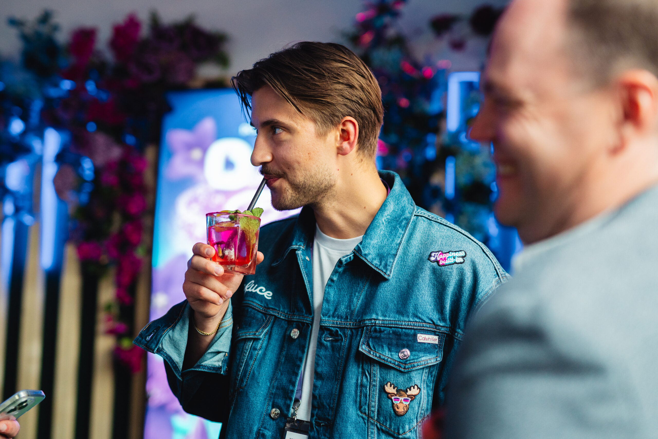 Gast drinkt een cocktail tijdens een event, met een verlichte bloemenwand op de achtergrond