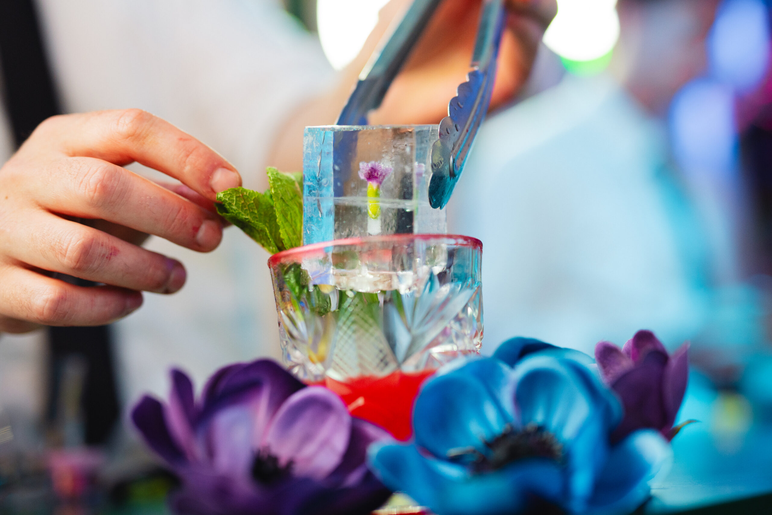 Bartender legt een ijsblok met een ingevroren bloem in een glas, met blauwe en paarse bloemen op de bar