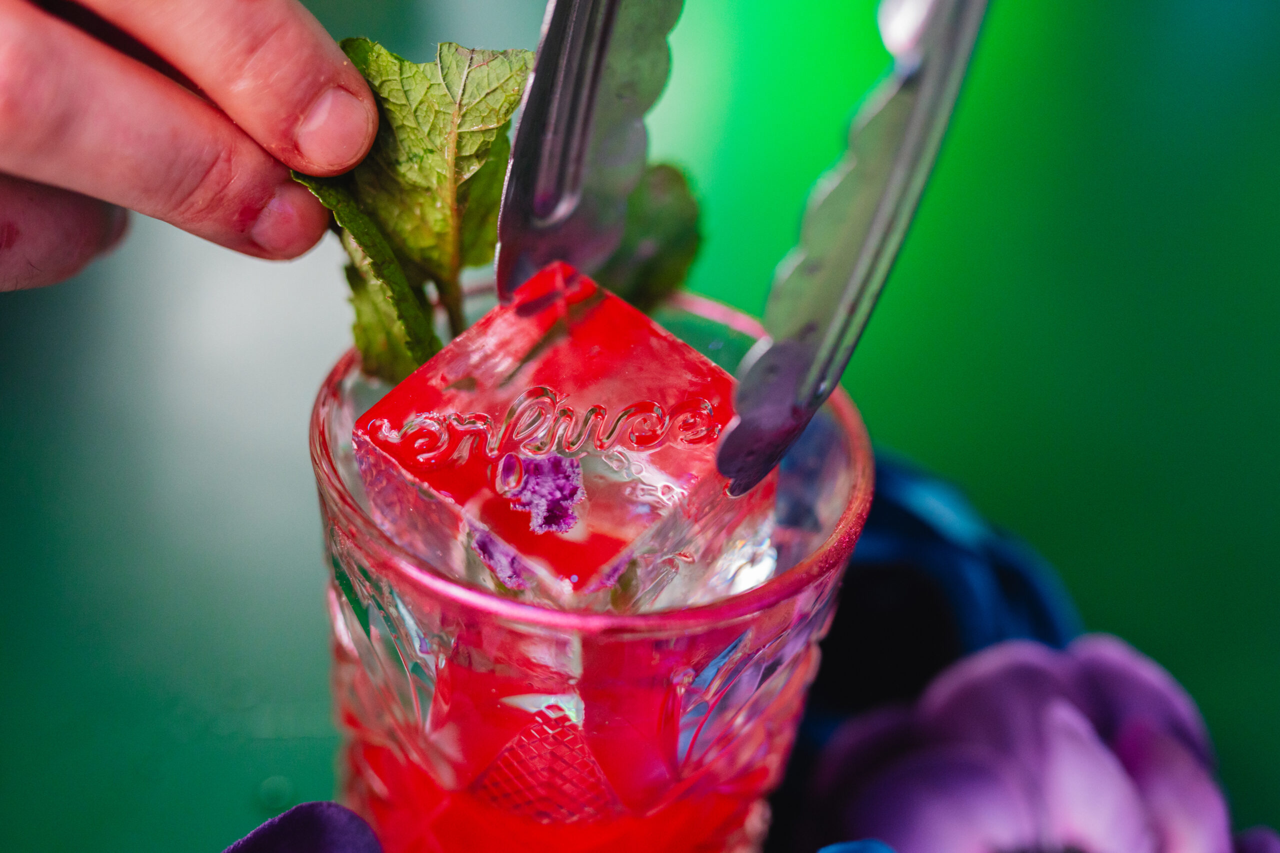 Bartender laat met een tang een rood ijsblok met ingegraveerde tekst en een paarse bloem in een cocktailglas zakken