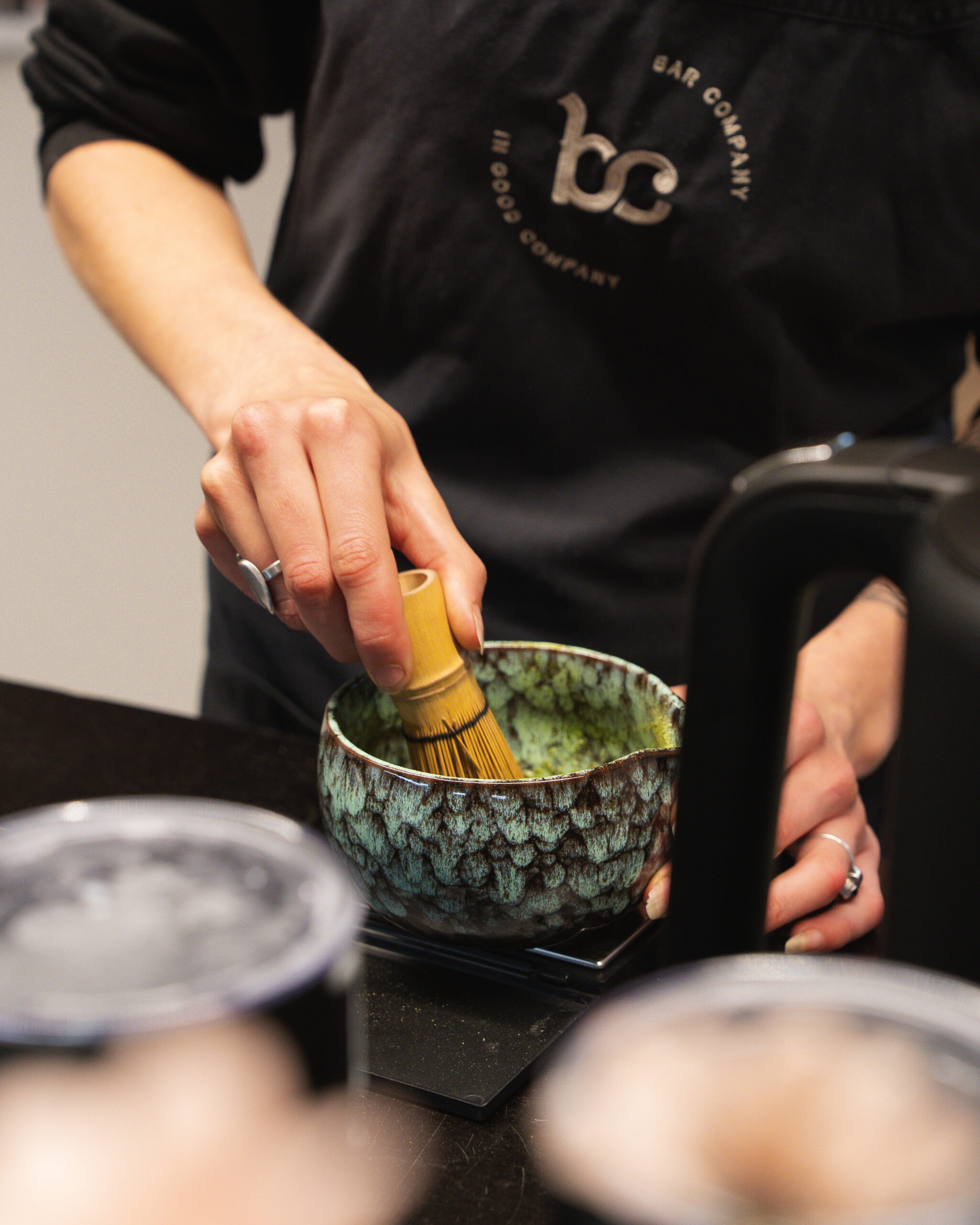 Barista klopt matcha met een bamboe garde in een groen gespikkelde kom, in schort van Bar Company