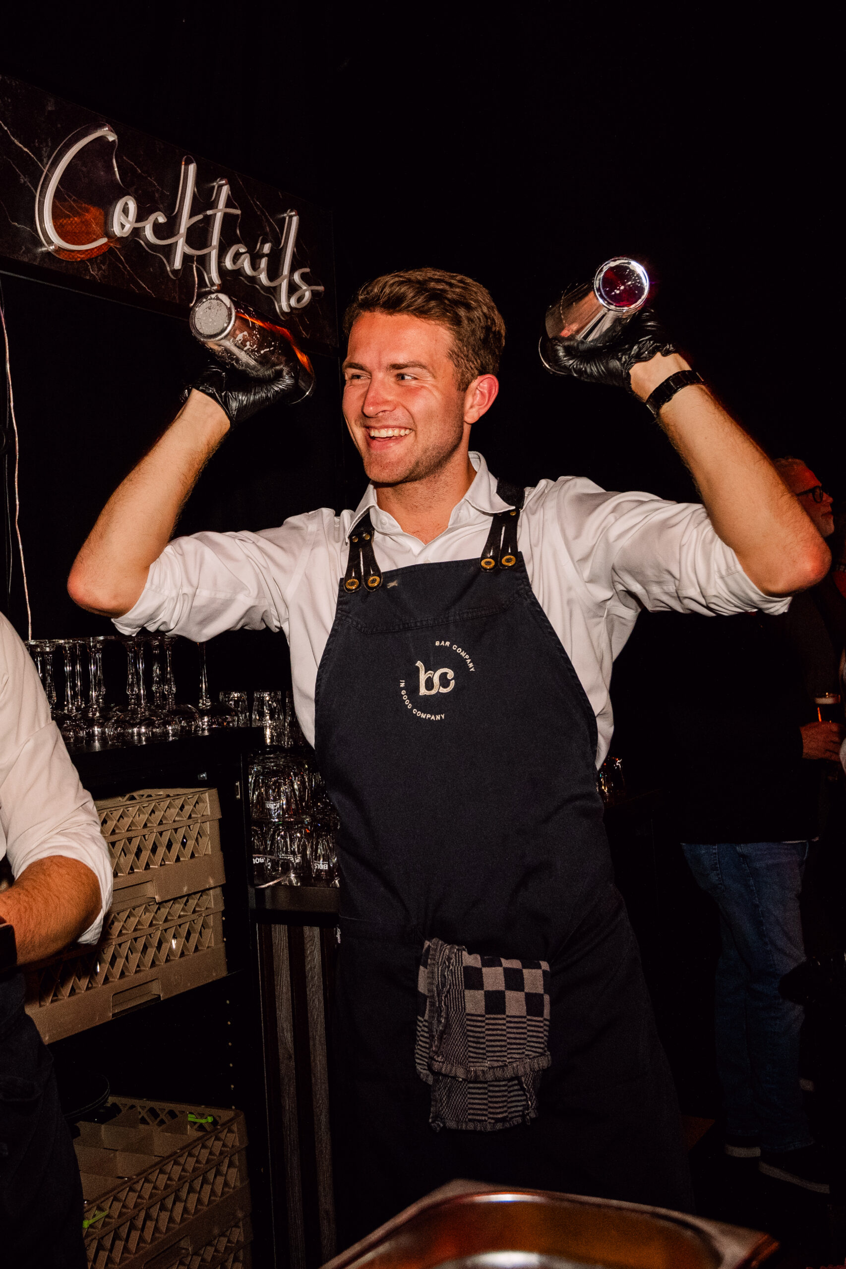 Lachende bartender van Bar Company houdt twee shakers omhoog onder een verlicht cocktailbord
