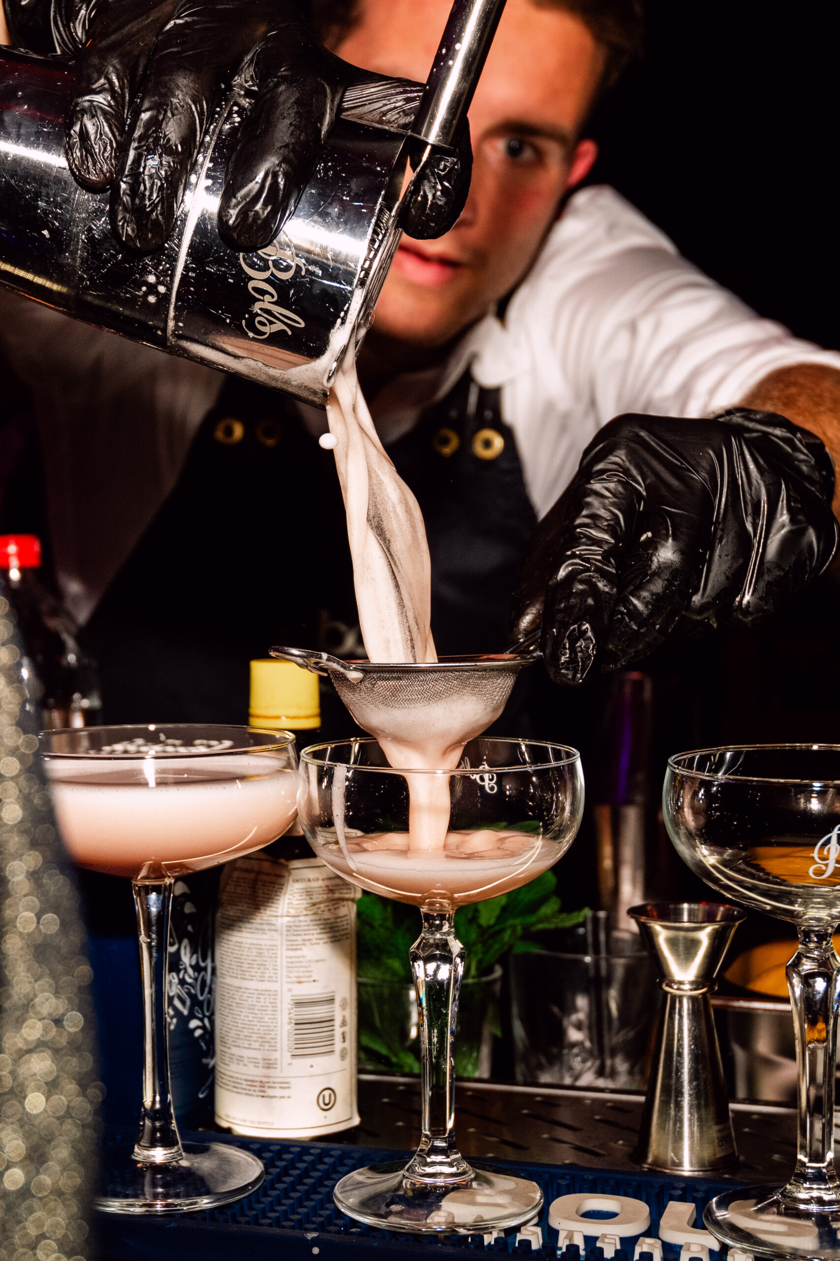 Bartender schenkt een roze cocktail door een zeefje in een coupe, met twee gevulde glazen ernaast
