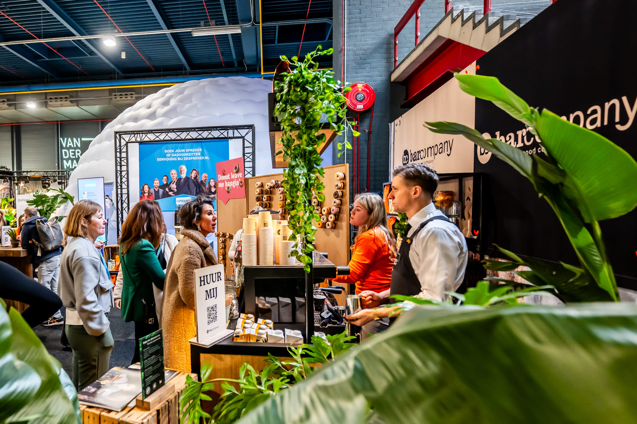 Bezoekers bij de Bar Company stand op een beurs, met bekers en apparatuur en medewerkers achter de bar.
