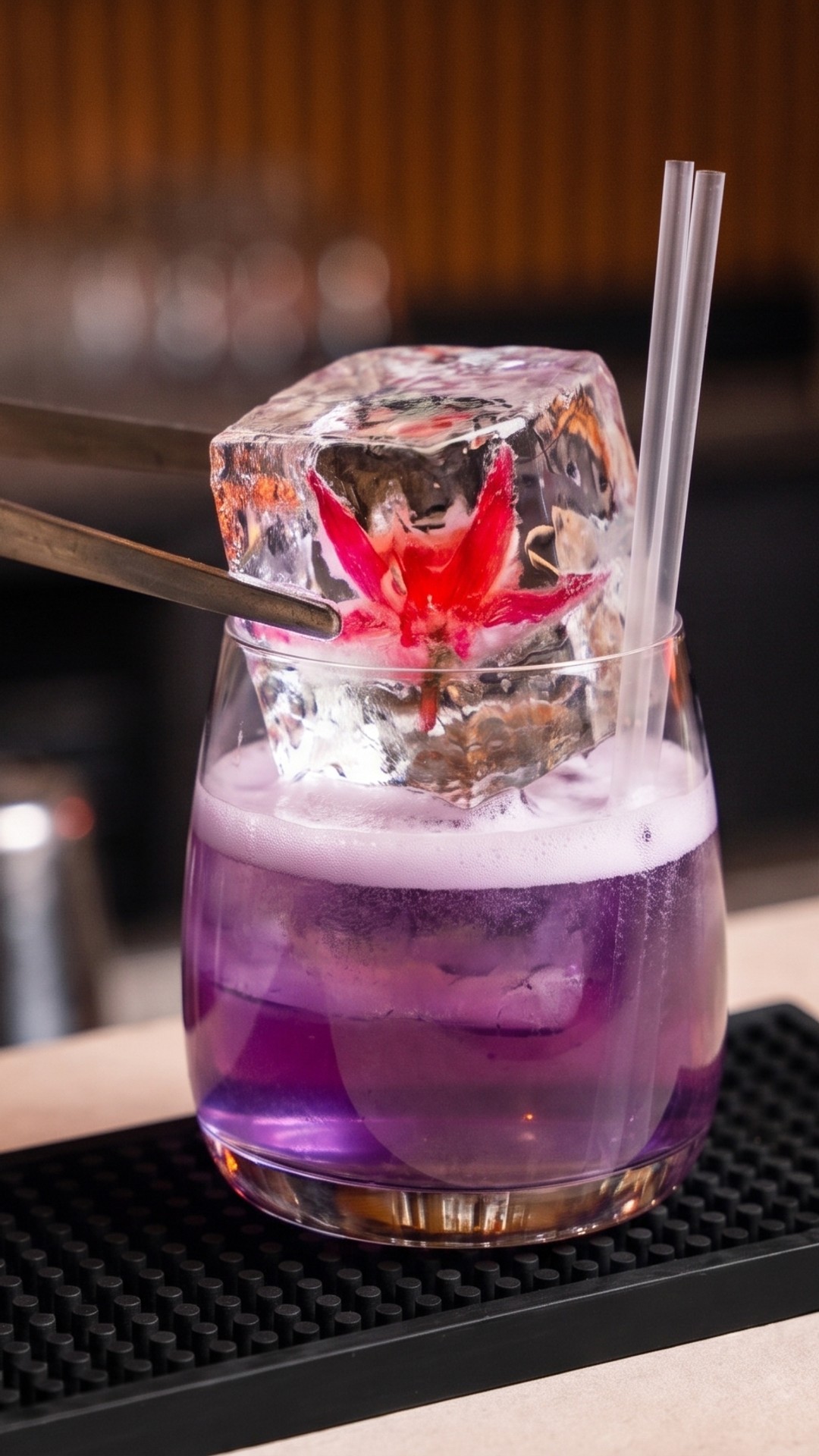 Large ice cube with a red flower frozen inside, attached to tongs, above a glass with a purple cocktail and straw.