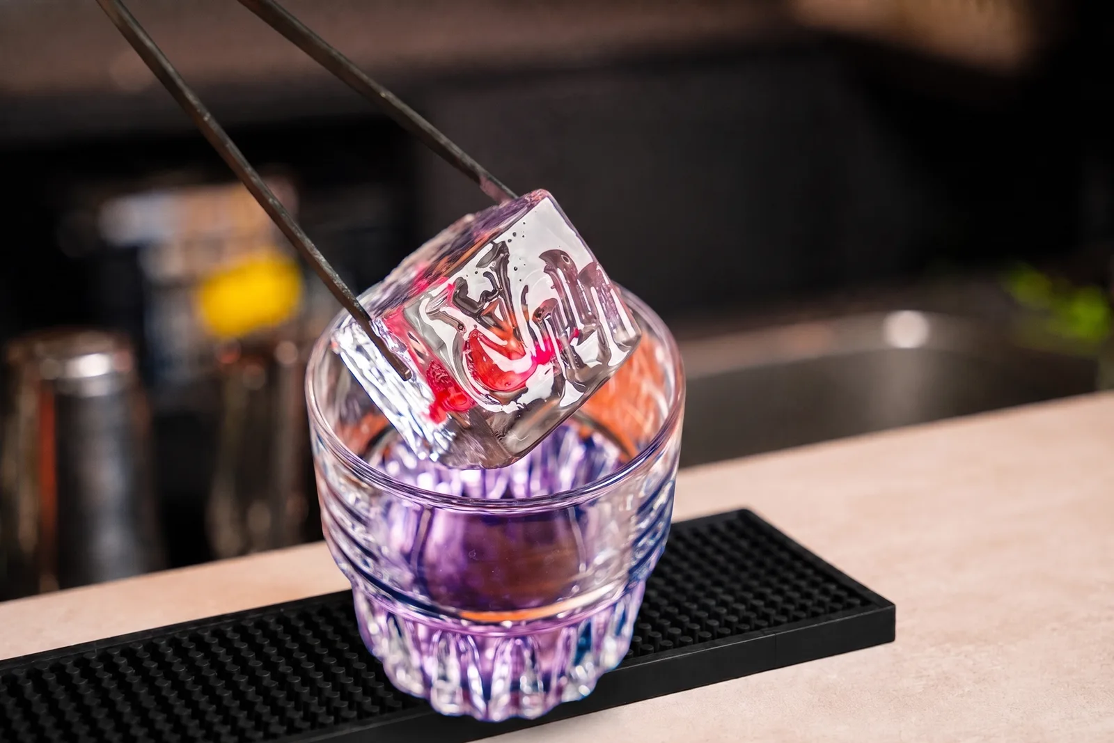 The bartender uses tongs to drop an ice cube with an engraved logo into a glass of purple cocktail.