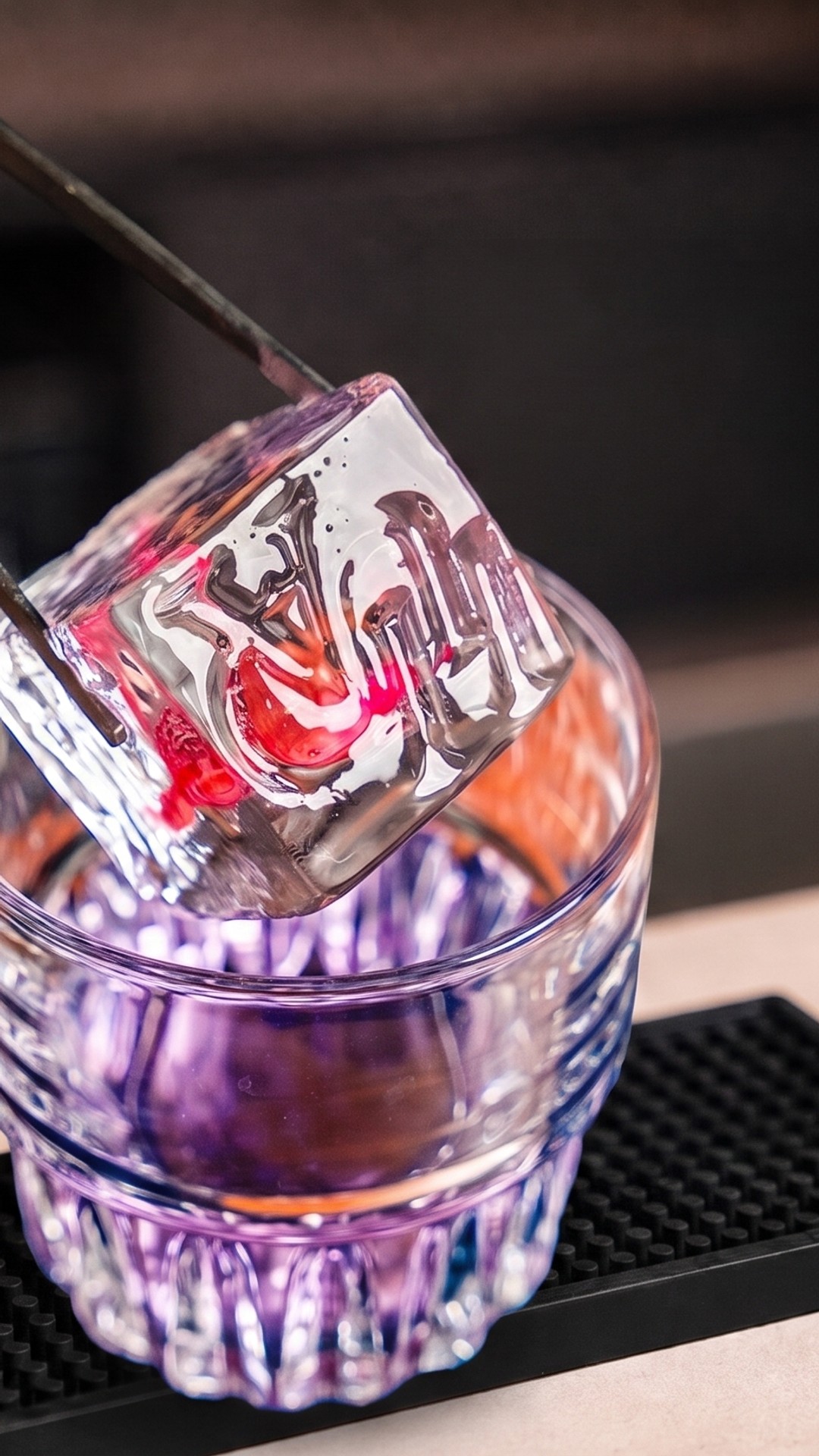 Ice cube with an engraved logo in red on tongs, just above a glass with a purple cocktail