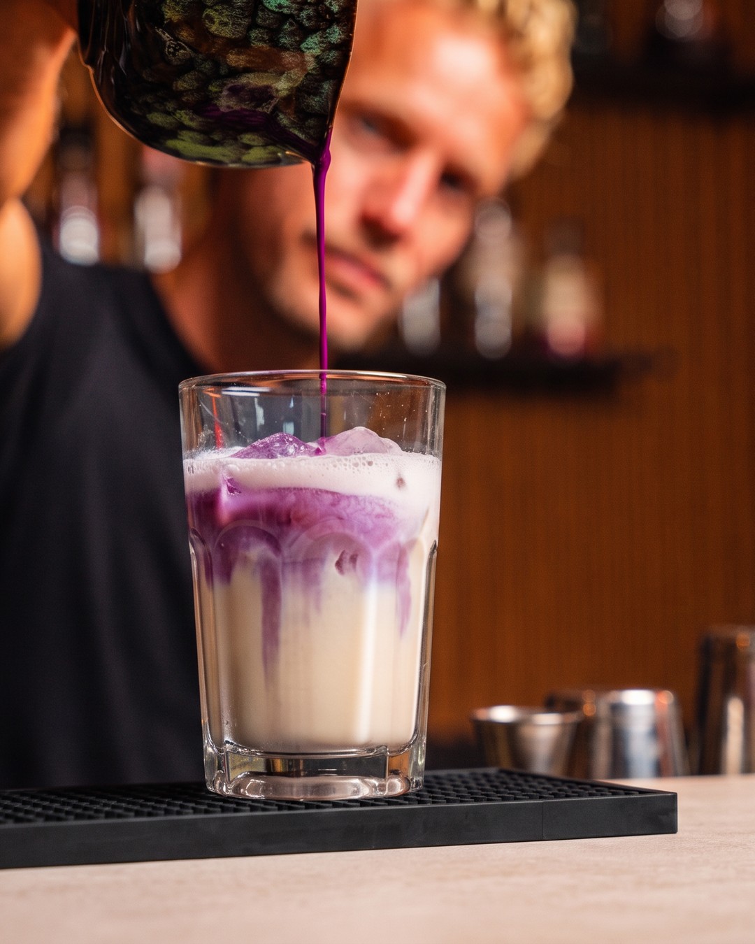 Barista schenkt paarse ube uit een kannetje in een hoog glas met melk en ijs op de bar
