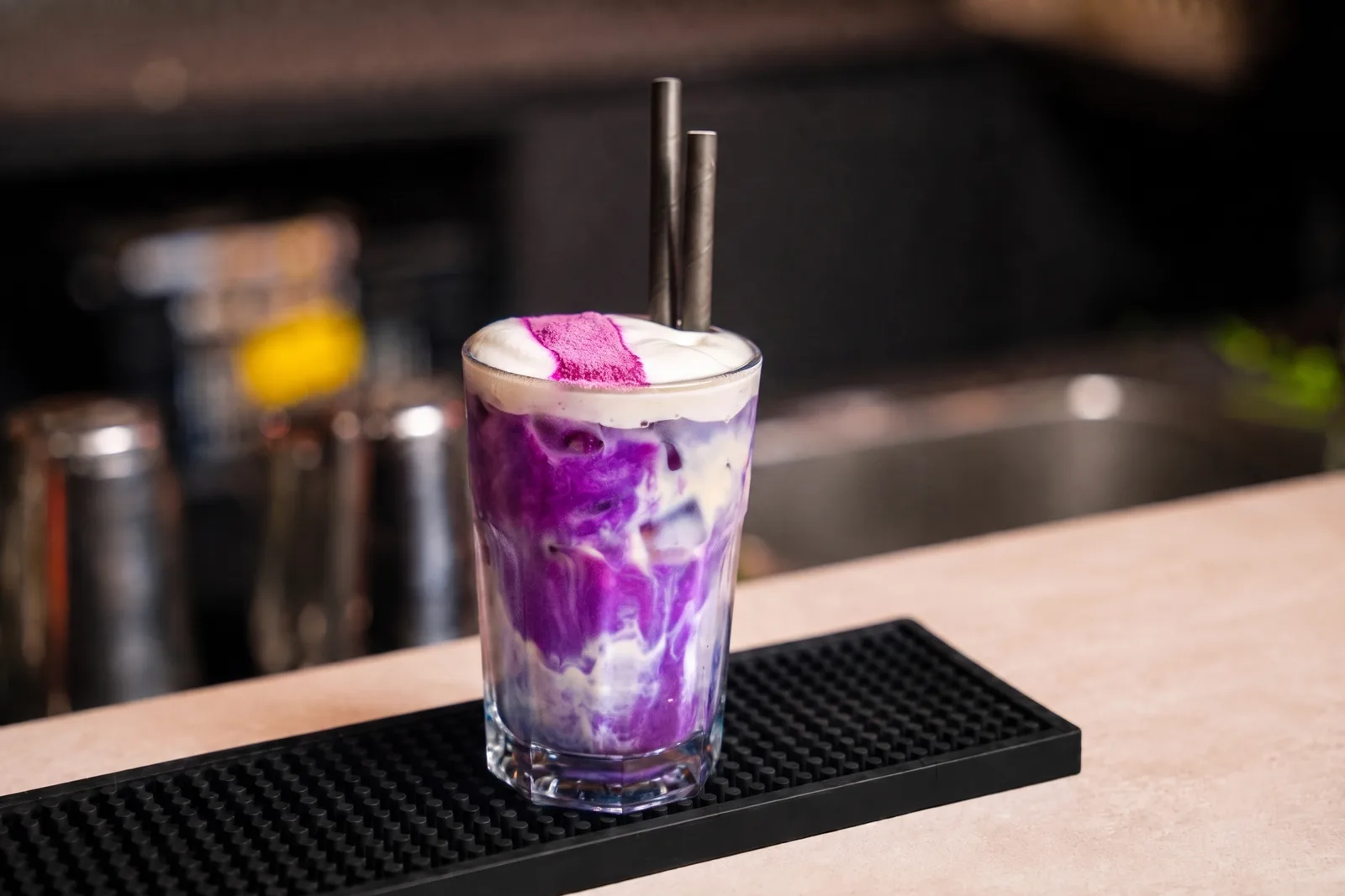 Glas ube met paarse en witte lagen door elkaar gemarmerd, wit schuim met paars poeder en twee zwarte rietjes op een barmat