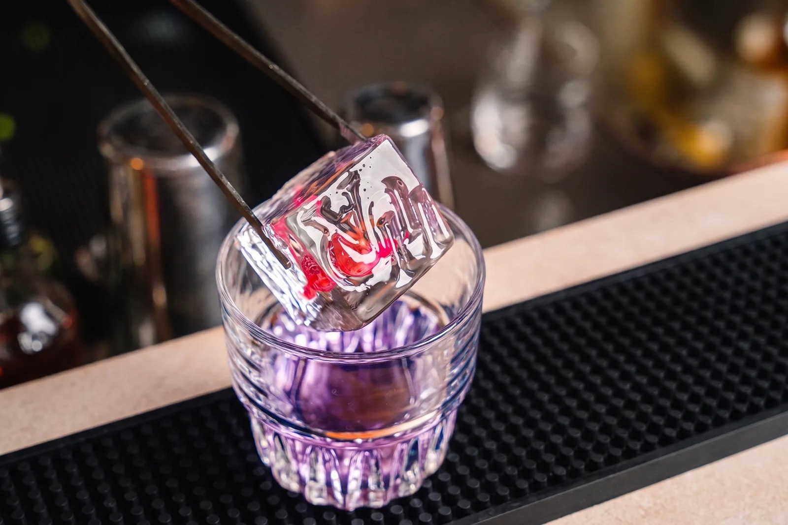 Using tongs, the bartender lowers an ice cube with a red-coloured engraved logo into a glass containing a purple cocktail