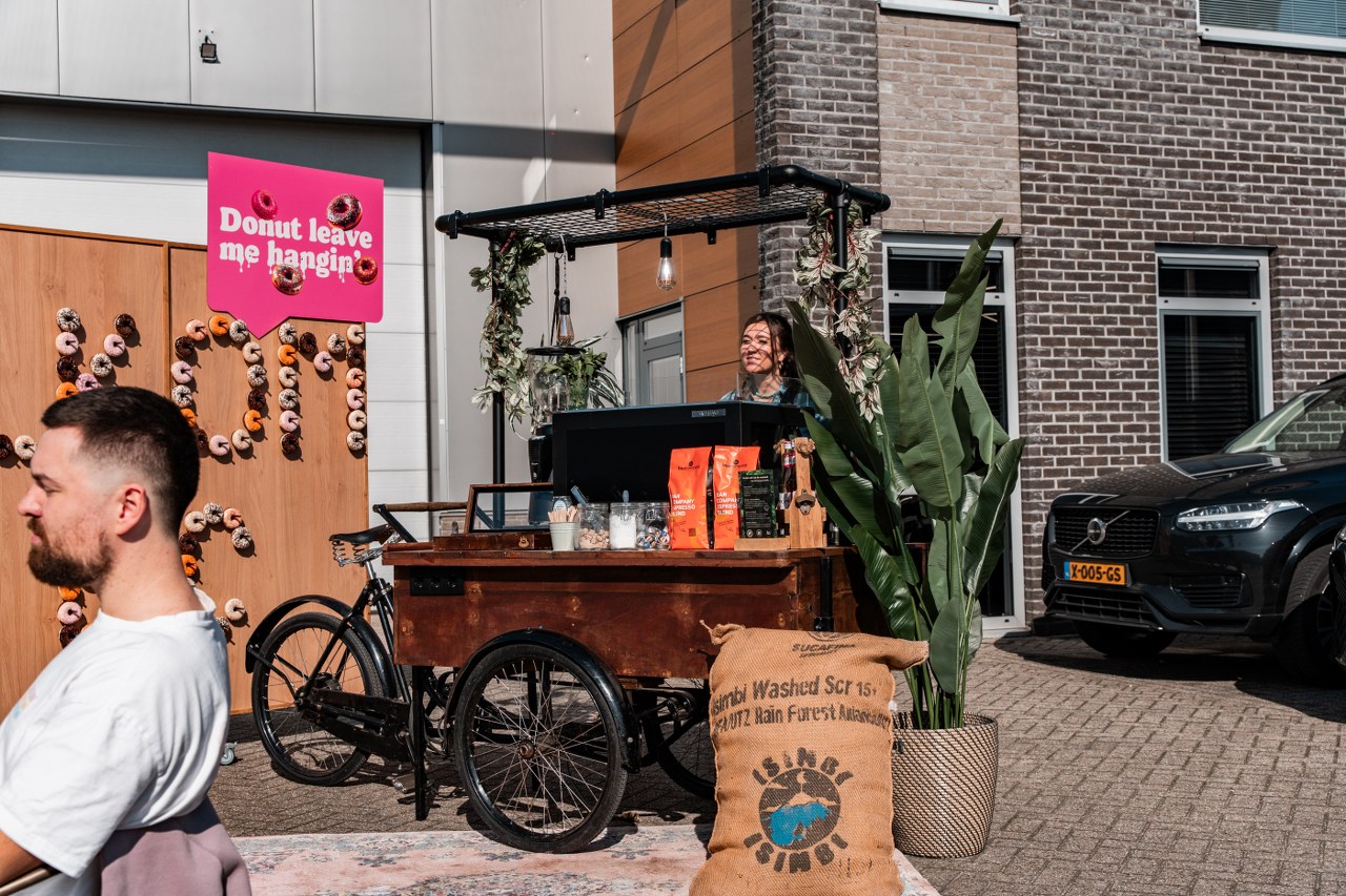 Barista achter een houten koffiefiets met hanglampjes en planten buiten, met een Donut Wall op de achtergrond.